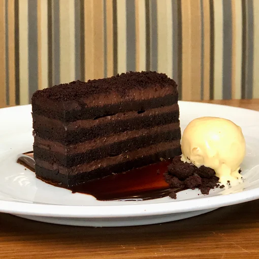 A slice of layered chocolate cake sits on a white plate with chocolate sauce pooling underneath, accompanied by a scoop of vanilla ice cream and crumbs, against a striped background.