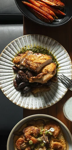 A plated roasted chicken breast sits over mushrooms and sauce on a patterned dish, with roasted carrots in a separate bowl and another dish partially visible on a wooden table.