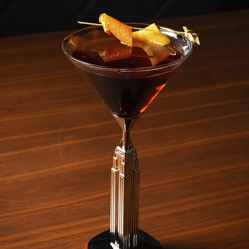 A dark brown cocktail sits in a tall martini glass with a decorative metallic stem, garnished with a skewered orange peel, placed on a wooden surface against a dark background.
