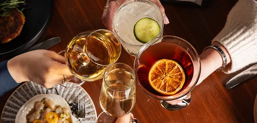our hands hold glasses together in a toast over a wooden table, including a white wine, a clear drink with cucumber slice, a dark cocktail with dried orange, and a tall glass of water.