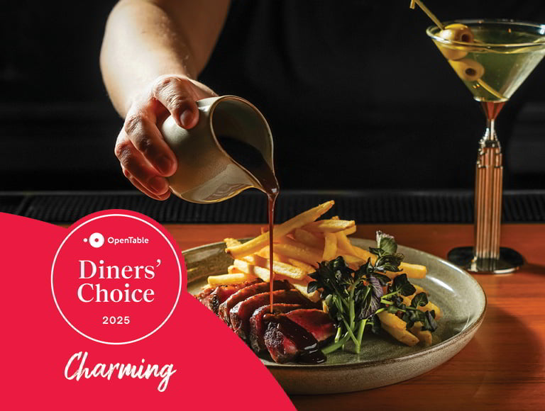 A hand pouring brown sauce over sliced steak served with French fries and greens, with a cocktail glass and “OpenTable Diners’ Choice 2025 Charming” logo visible.