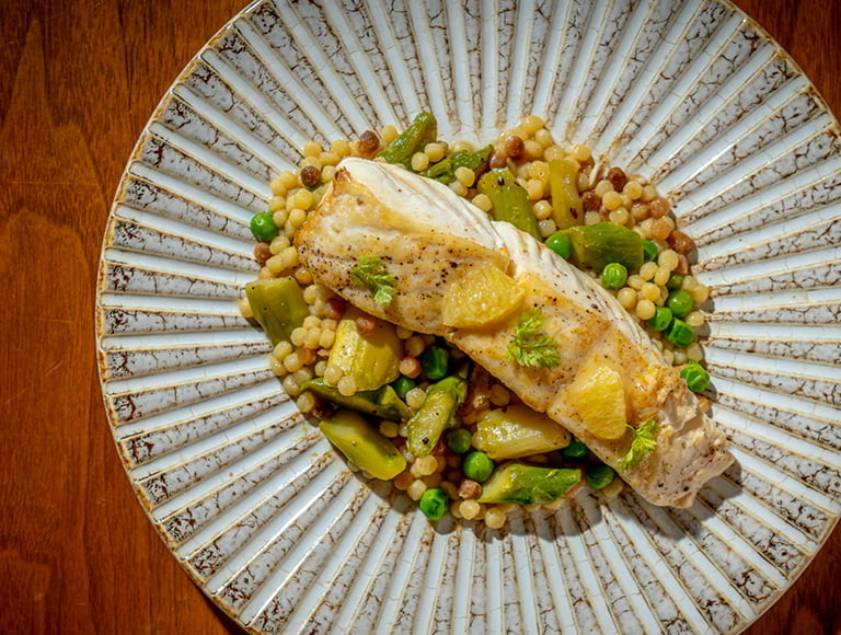 A grilled white fish fillet topped with herbs and citrus pieces, served over pearl couscous with peas and green vegetables on a patterned ceramic plate.