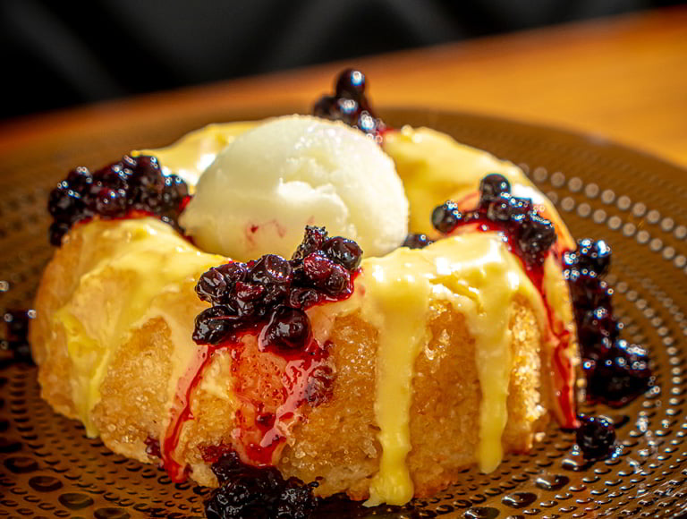 A round sponge dessert drizzled with yellow sauce and topped with dark berries and a scoop of white ice cream on a brown plate.